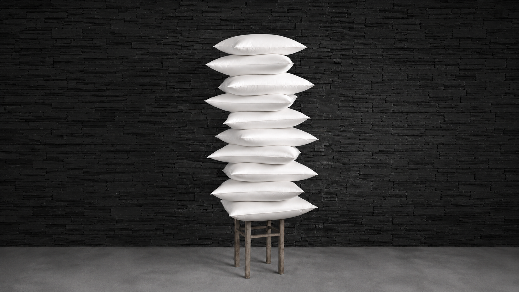 Tall stack of white pillows balanced on a wooden stool against a dark stone wall