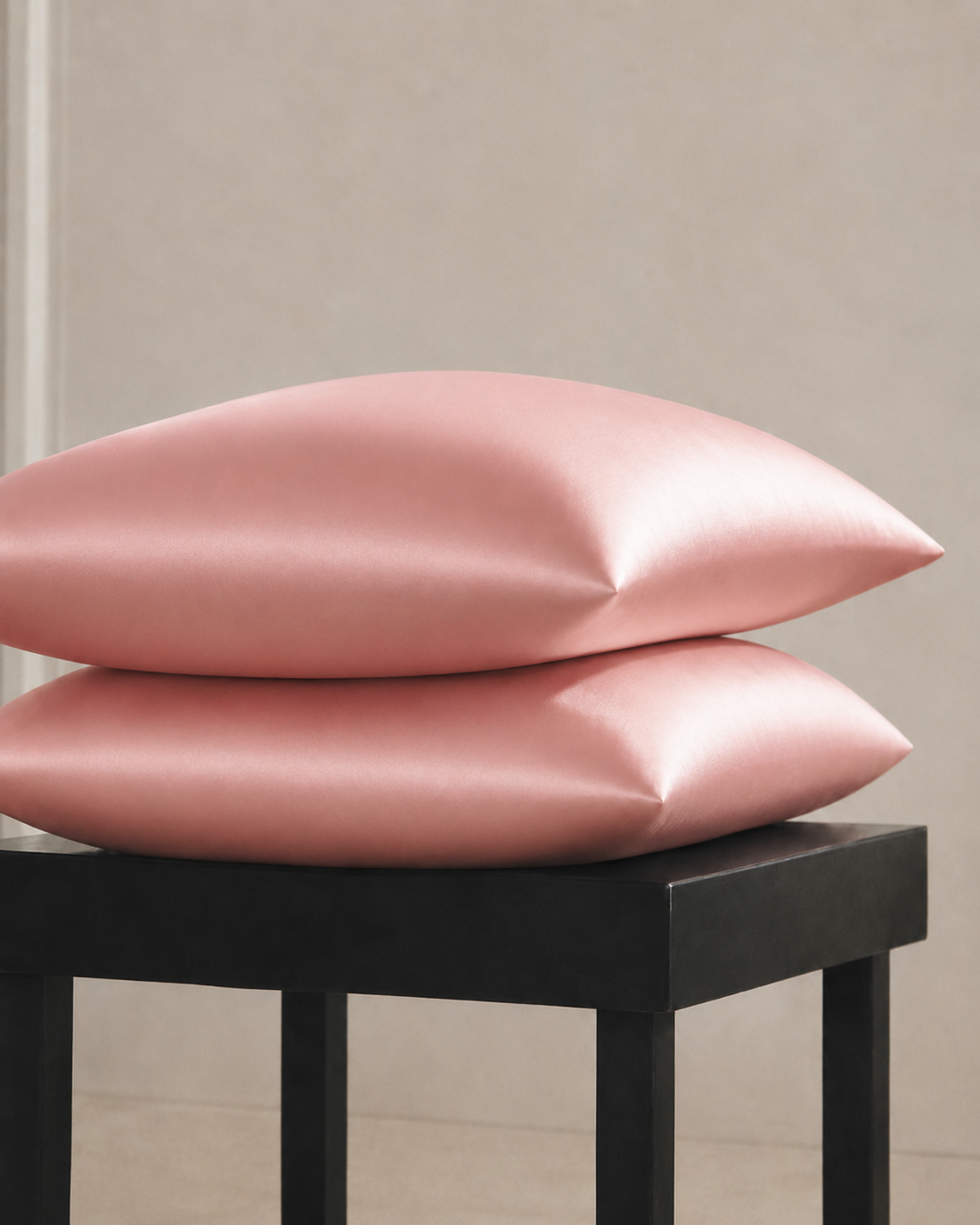 Two Blush Pink silk pillowcases stacked on a black stool against a pale wall