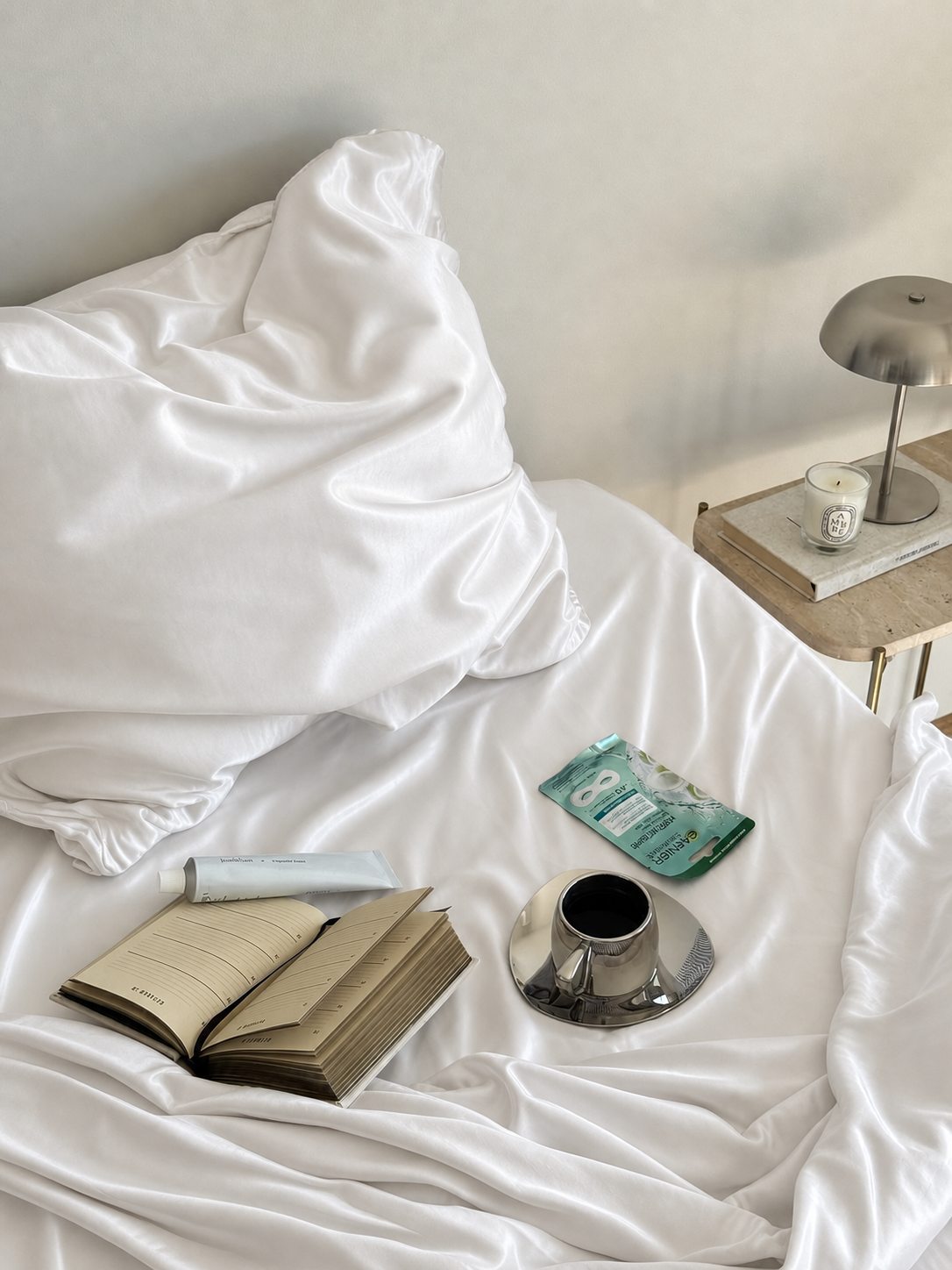 White silk bedding rumpled on a bed with a notebook, coffee cup and skincare products